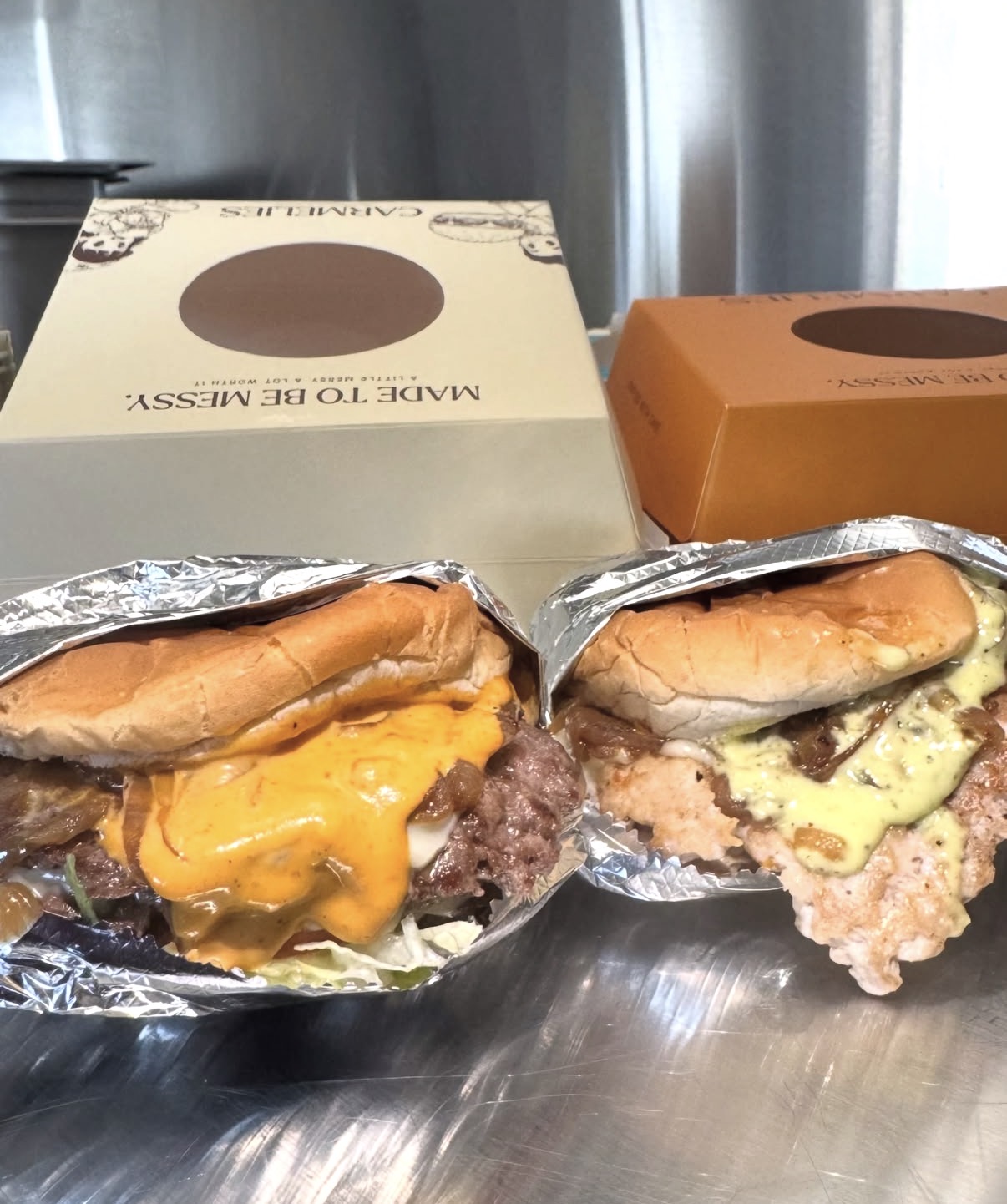 Beef and chicken smashburgers in foil side by side with Carmelies boxes behind them